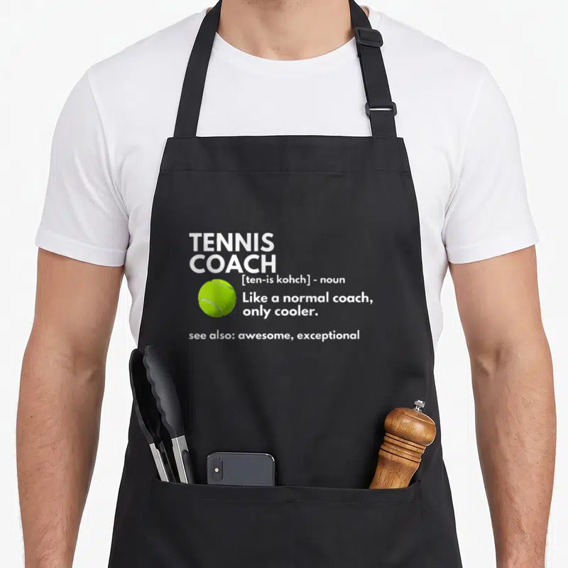 Coach tennis mom apron - Stacked white text defining tennis coach next to a neon yellow tennis ball — shown on a black cotton canvas apron against a white background