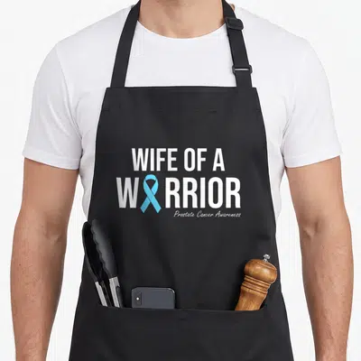Family wife apron - White text Wife of a Warrior with light blue ribbon replacing the O — shown on a black cotton canvas apron against a white background
