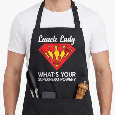 Superhero lunch lady apron - Red and yellow shield with four kitchen utensils and white text asking about superhero — shown on a black cotton canvas apron against a white background