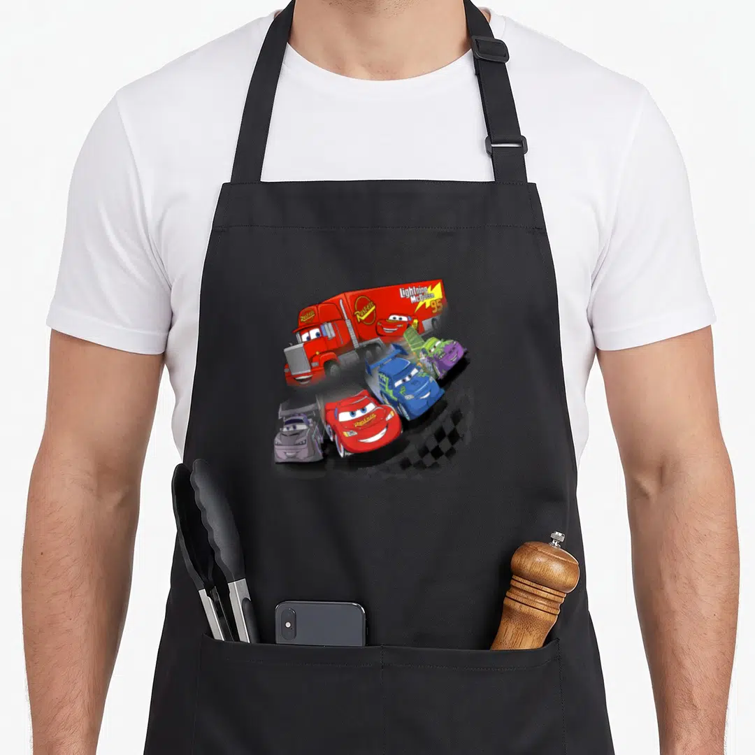 Car mac apron - Red race car and rival cars clustered near Mack truck with Rust-eze logo and checkered flag — shown on a black cotton canvas apron against a white background