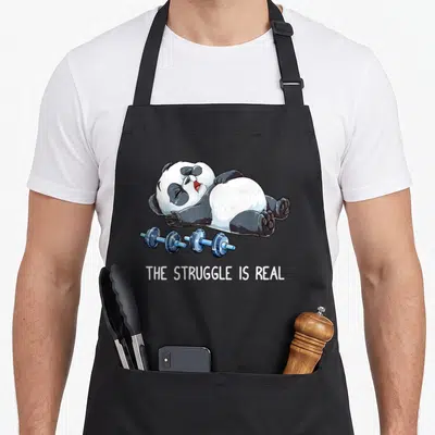 Struggle deadlift apron - Cartoon panda lying on back above blue dumbbells with text THE STRUGGLE IS REAL — shown on a black cotton canvas apron against a white background