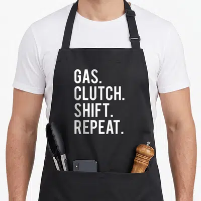 Gas car apron - White stacked text reading GAS CLUTCH SHIFT REPEAT on black fabric — shown on a black cotton canvas apron against a white background