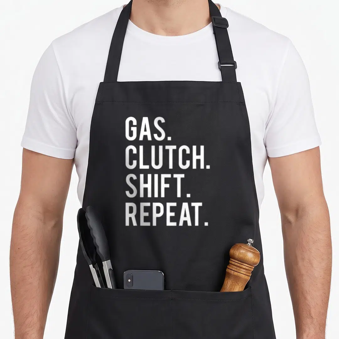 Gas car apron - White stacked text reading GAS CLUTCH SHIFT REPEAT on black fabric — shown on a black cotton canvas apron against a white background