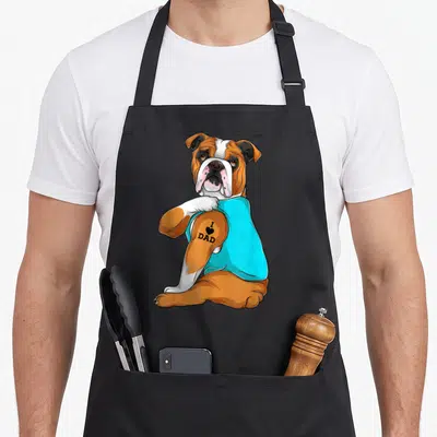 English tattoo aprons - Cartoon bulldog in cyan shirt with I Love Dad tattoo on brown arm — shown on a black cotton canvas apron against a white background