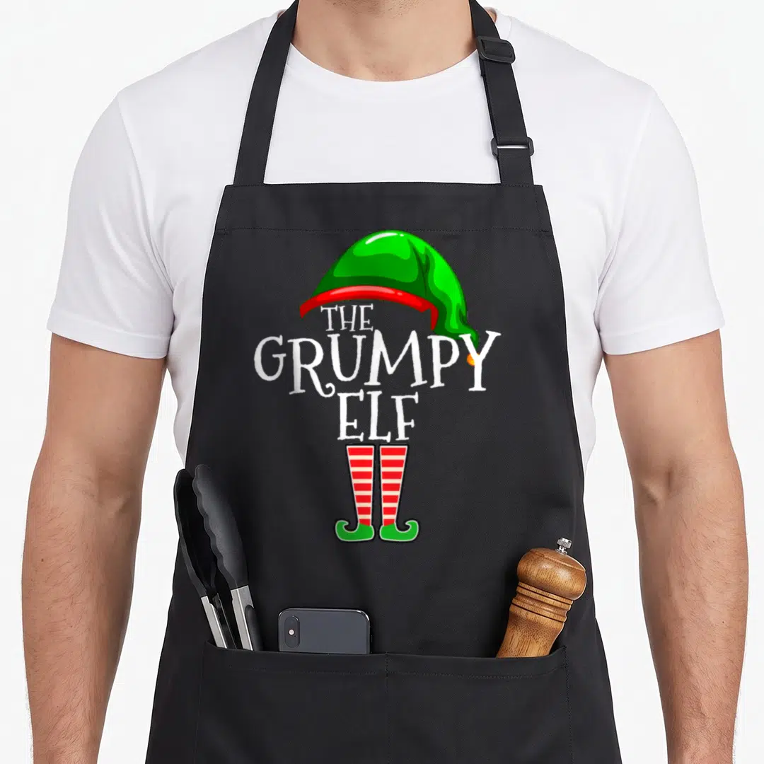 Grumpy christmas elf apron - Stacked white text between green hat and red striped elf shoes in cartoon style — shown on a black cotton canvas apron against a white background