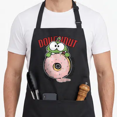 Cut om symbol apron - Green monster holding pink frosted donut with DOUGHNUT and DONUT NOMMIE text — shown on a black cotton canvas apron against a white background