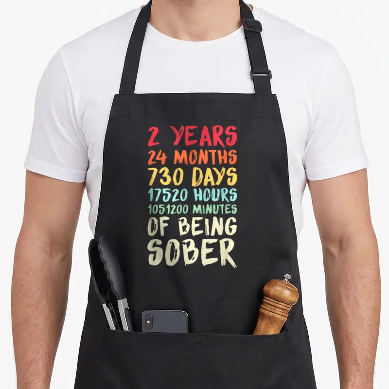 Recovery clean apron - Stacked colorful text listing 2 years, 24 months, 730 days, hours and minutes of being sober — shown on a black cotton canvas apron against a white background