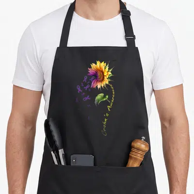 Crohn sunflower apron - Watercolor sunflower with purple ribbons, green leaf, and curved 'Crohn's Awareness' text — shown on a black cotton canvas apron against a white background