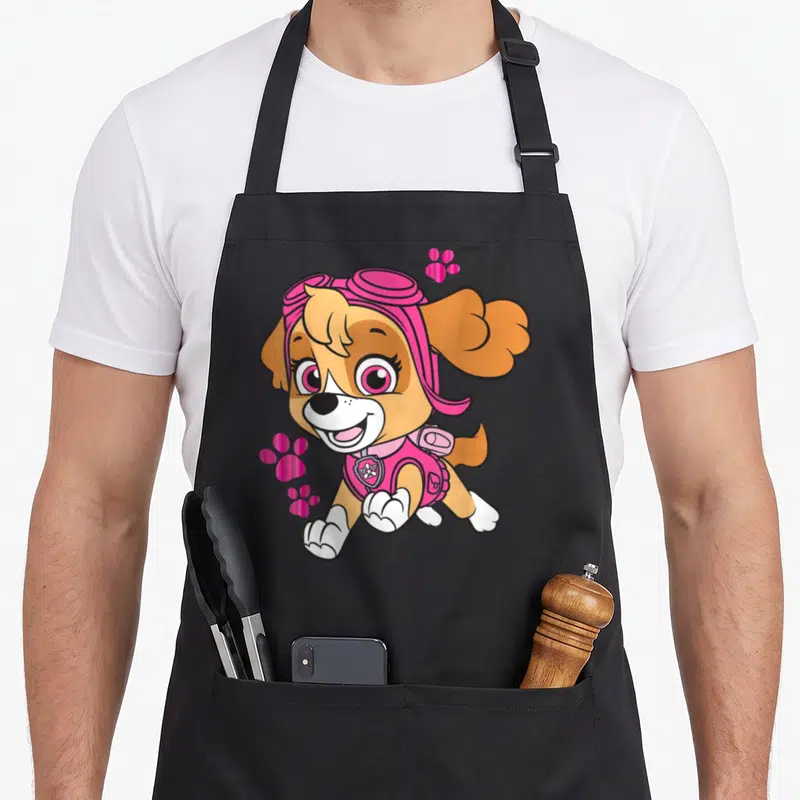 Skye novelty apron - Cartoon dog in pink goggles and vest with tan fur and pink paw prints — shown on a black cotton canvas apron against a white background