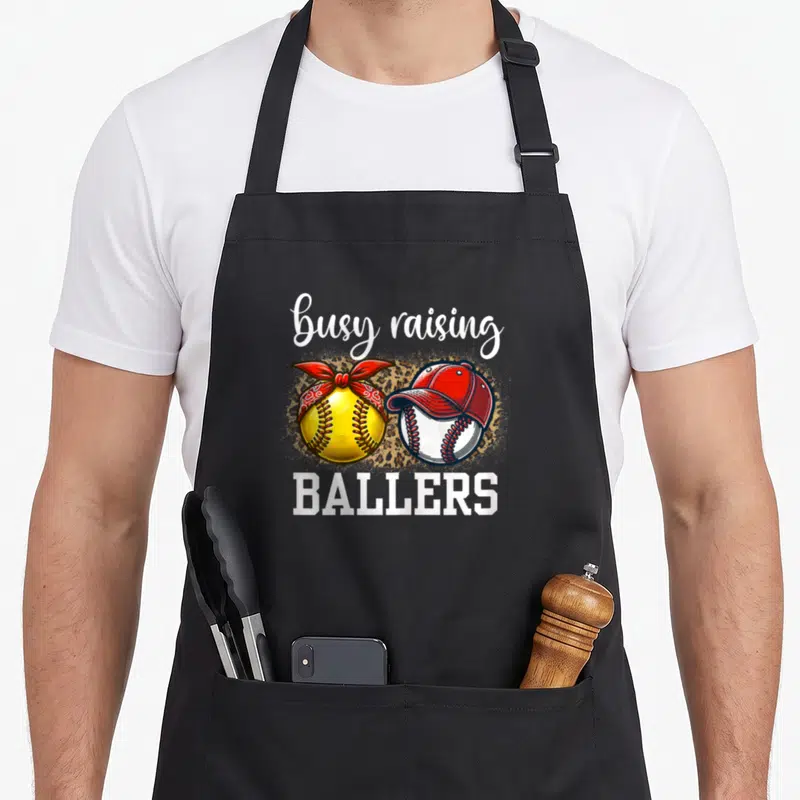 Busy leopard print apron - busy raising ballers text with softball bandana and baseball cap over leopard print — shown on a black cotton canvas apron against a white background