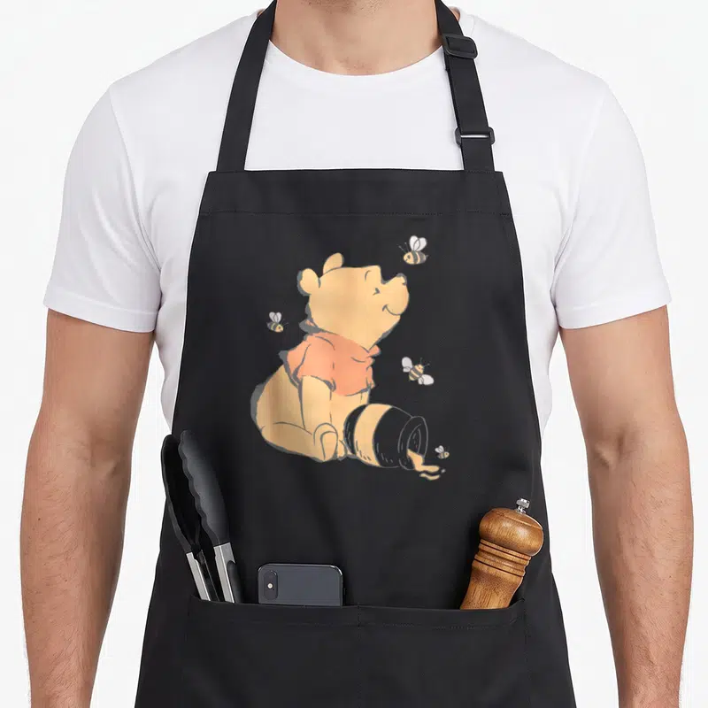 Funny honey bee apron - Cartoon bear in red shirt sitting next to tipped pot with four bees on grey background — shown on a black cotton canvas apron against a white background