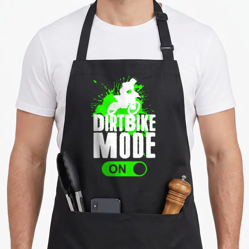 Mode bike apron - White dirt bike rider silhouette on green splatter with DIRTBIKE MODE ON text and toggle switch — shown on a black cotton canvas apron against a white background