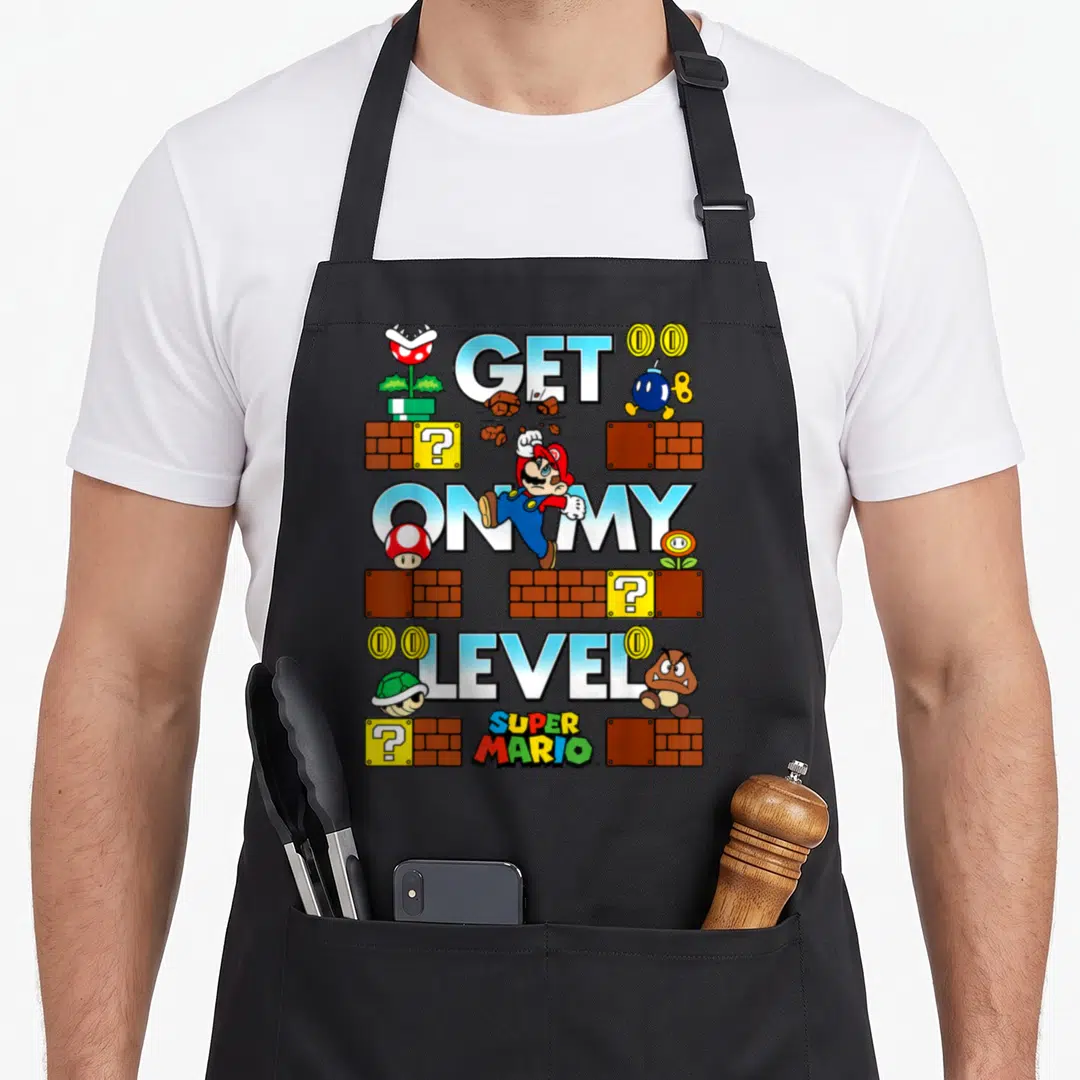 Level game aprons - MARIO jumping through stacked GET ON MY LEVEL text with bricks, coins, mushrooms, and enemies — shown on a black cotton canvas apron against a white background