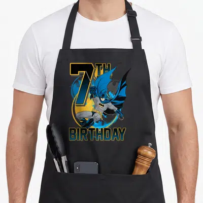 7th action birthday apron - Cartoon Batman leaping in blue and yellow before 7TH BIRTHDAY text — shown on a black cotton canvas apron against a white background