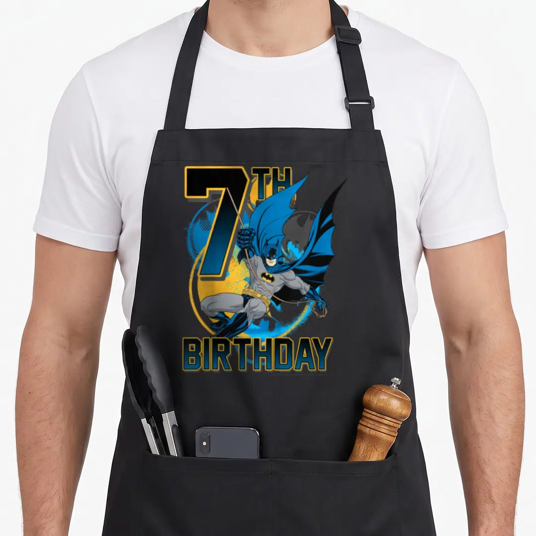 7th action birthday apron - Cartoon Batman leaping in blue and yellow before 7TH BIRTHDAY text — shown on a black cotton canvas apron against a white background
