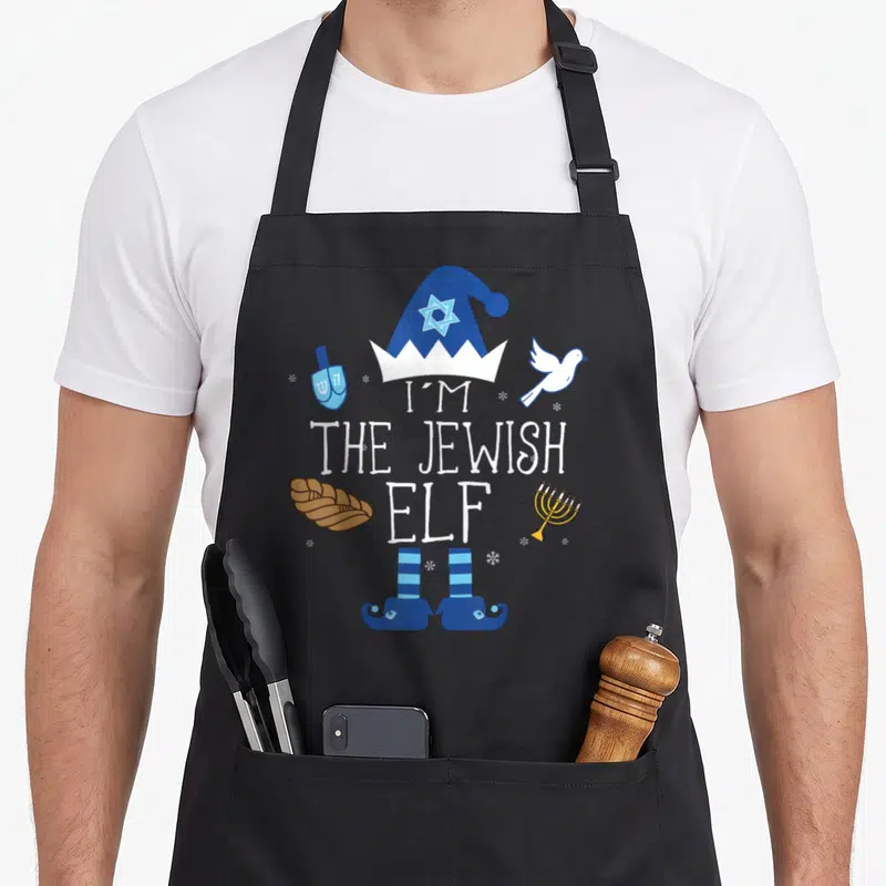 Hanukkah jewish apron - Cartoon elf hat and shoes framing 'I'M THE JEWISH ELF' text with menorah, challah, dreidel — shown on a black cotton canvas apron against a white background