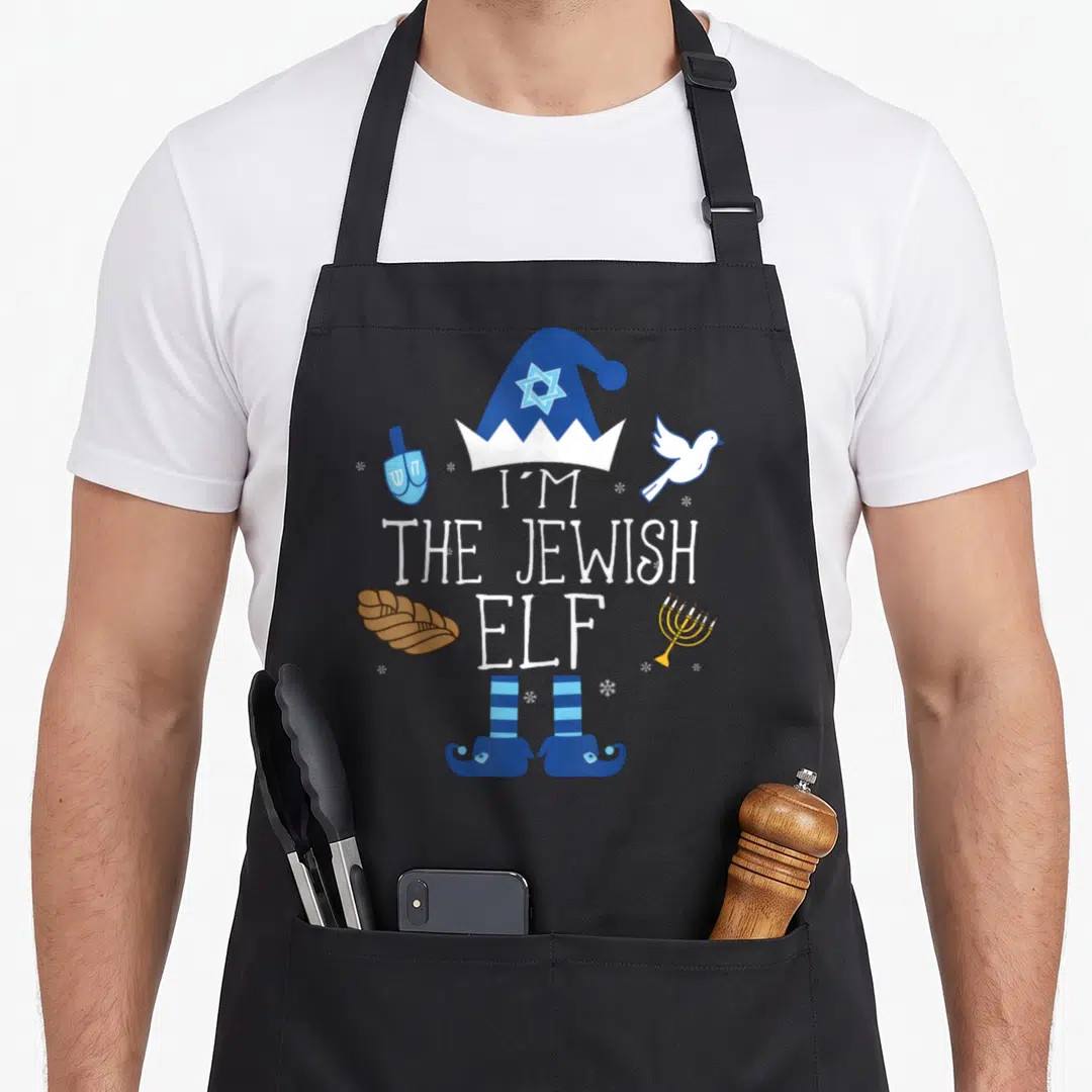 Hanukkah jewish apron - Cartoon elf hat and shoes framing 'I'M THE JEWISH ELF' text with menorah, challah, dreidel — shown on a black cotton canvas apron against a white background