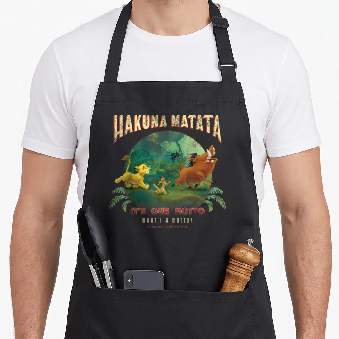Motto lion apron - Cartoon lion cub and text Hakuna Matata It's Our Motto inside a green jungle circle — shown on a black cotton canvas apron against a white background