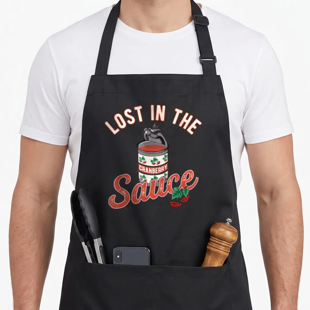 Lost thanksgiving day aprons - Cartoon can of cranberry sauce with text LOST IN THE CRANBERRY Sauce and berry cluster — shown on a black cotton canvas apron against a white background