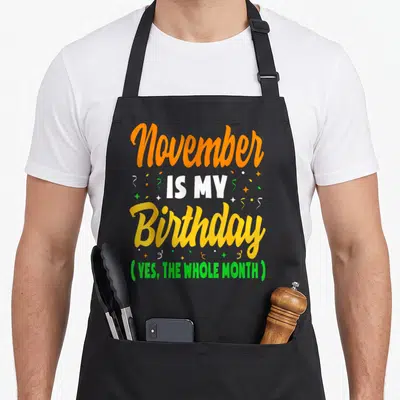 November whole birthday apron - Stacked orange yellow green and white text reading November IS MY Birthday YES — shown on a black cotton canvas apron against a white background