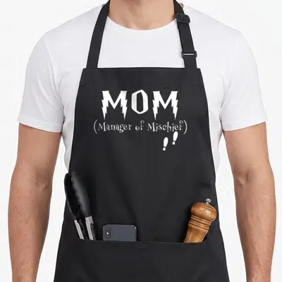 Manager production quotes apron - White stacked text reading MOM Manager of Mischief with two exclamation marks below — shown on a black cotton canvas apron against a white background