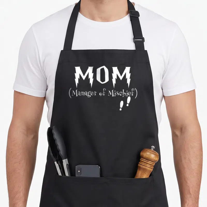 Manager production quotes apron - White stacked text reading MOM Manager of Mischief with two exclamation marks below — shown on a black cotton canvas apron against a white background