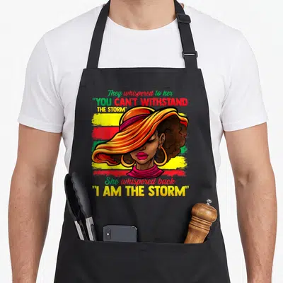 Proud african american aprons - Woman with I Am The Storm text over yellow red green stripes — shown on a black cotton canvas apron against a white background