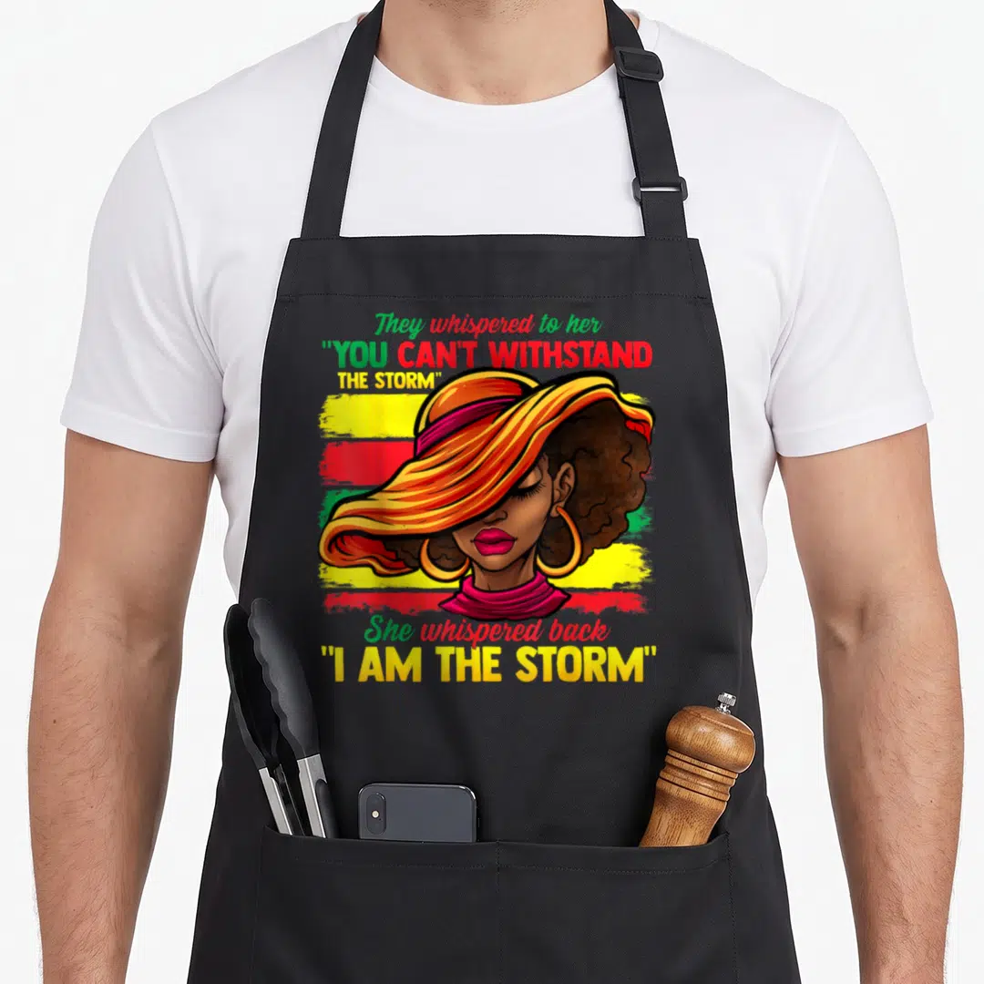 Proud african american aprons - Woman with I Am The Storm text over yellow red green stripes — shown on a black cotton canvas apron against a white background