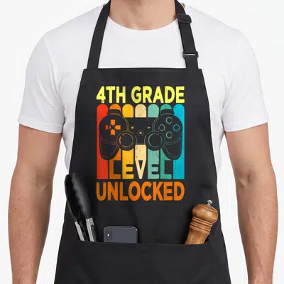 Hello 4th video game apron - Grey game controller silhouette over five colorful vertical stripes with 4TH GRADE text — shown on a black cotton canvas apron against a white background