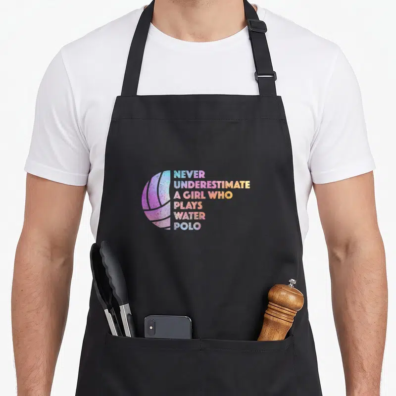 Athlete water polo apron - Colorful water polo ball next to stacked text reading never underestimate a girl who plays — shown on a black cotton canvas apron against a white background