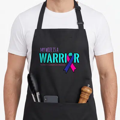 Thyroid wife apron - My Wife Is A Warrior text with pink and blue awareness ribbon — shown on a black cotton canvas apron against a white background