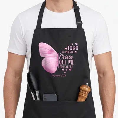 Christian spanish aprons - Pink butterfly next to Todo Lo Puedo En Cristo Que Me Fortalece text and small hearts — shown on a black cotton canvas apron against a white background