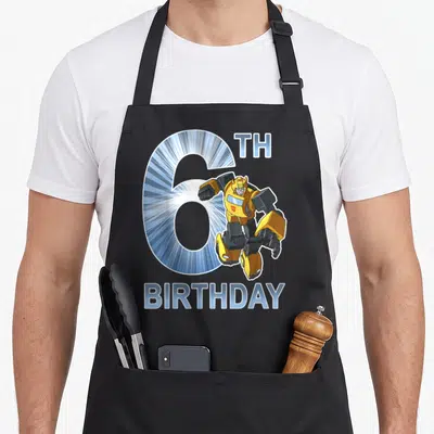 Bumblebee 6th birthday apron - Cartoon Bumblebee robot overlapping a large blue number 6 with TH BIRTHDAY text — shown on a black cotton canvas apron against a white background