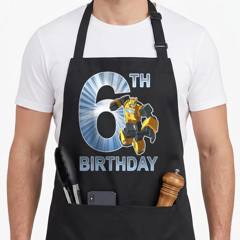 Bumblebee 6th birthday apron - Cartoon Bumblebee robot overlapping a large blue number 6 with TH BIRTHDAY text — shown on a black cotton canvas apron against a white background