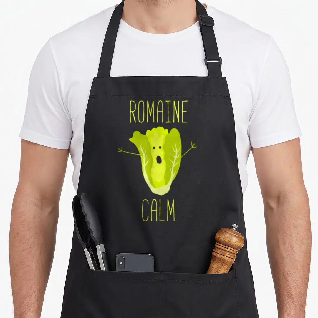 Pun gardener apron - Green cartoon romaine lettuce with arms and Romaine Calm text — shown on a black cotton canvas apron against a white background