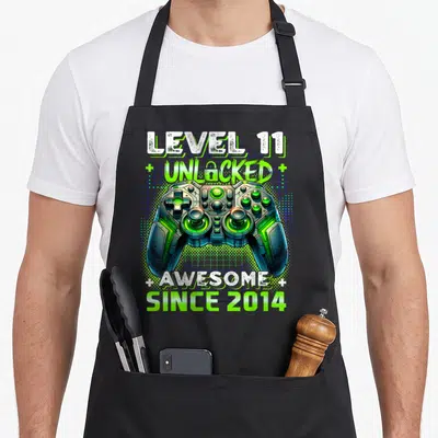 11th birthday level video game apron - Game controller with LEVEL 11 UNLOCKED AWESOME SINCE 2014 text and blue grid — shown on a black cotton canvas apron against a white background