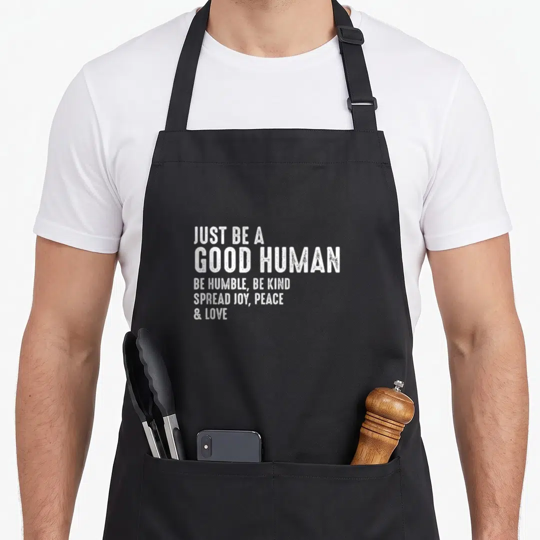 Human spread apron - White stacked text reading JUST BE A GOOD HUMAN BE HUMBLE BE KIND SPREAD JOY PEACE & LOVE — shown on a black cotton canvas apron against a white background