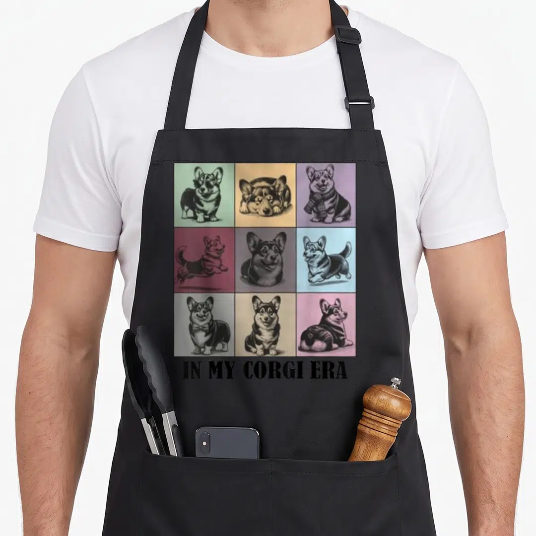 Corgi era dog apron - Nine cartoon corgis in colored squares above bold text reading IN MY CORGI ERA — shown on a black cotton canvas apron against a white background