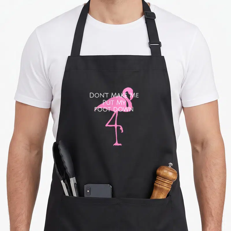 Cute awareness pink flamingo apron - Pink flamingo silhouette with white text DON'T MAKE ME PUT MY FOOT DOWN — shown on a black cotton canvas apron against a white background