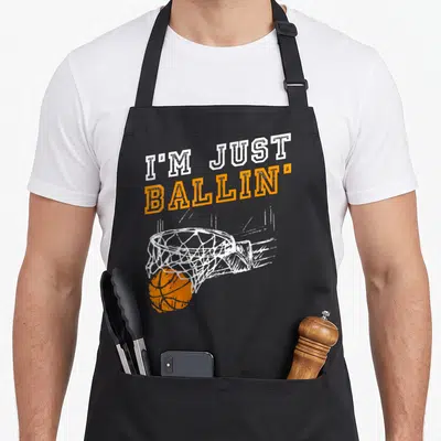 Youth basketball coach apron - I'M JUST BALLIN text over distressed orange basketball going through white hoop net — shown on a black cotton canvas apron against a white background