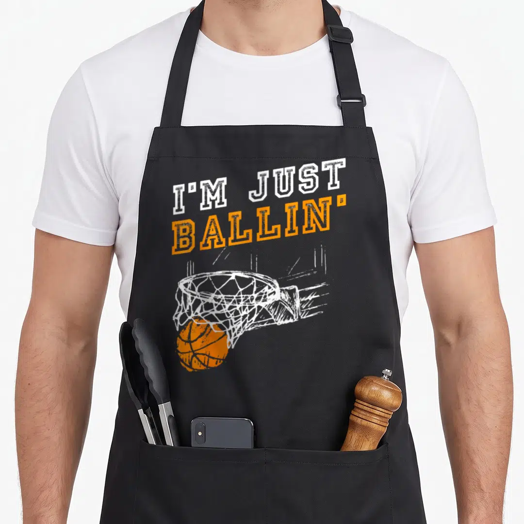 Youth basketball coach apron - I'M JUST BALLIN text over distressed orange basketball going through white hoop net — shown on a black cotton canvas apron against a white background