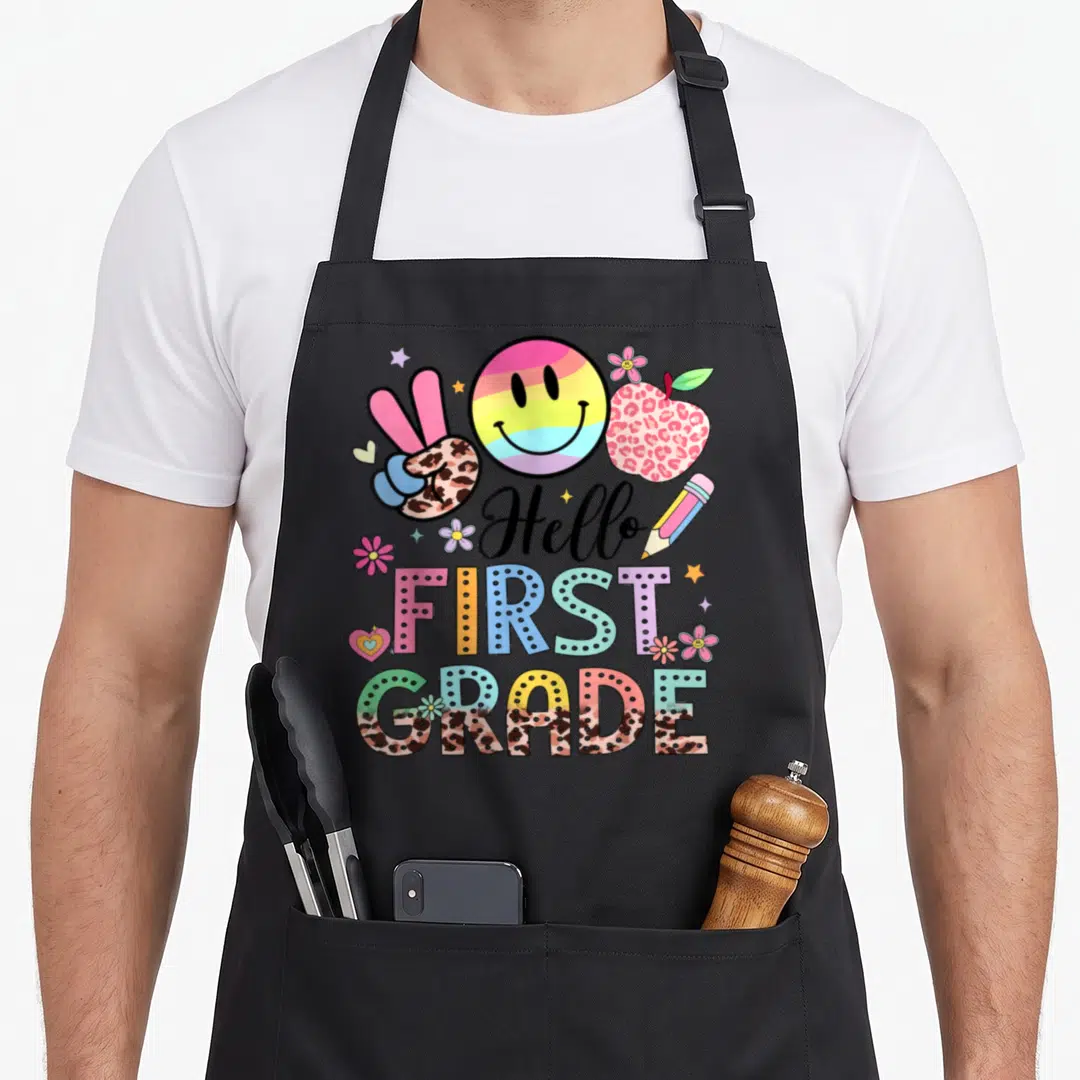 Hello 1st art teacher aprons - Rainbow smiley, leopard apple, peace hand, Hello FIRST GRADE text with stars and flowers — shown on a black cotton canvas apron against a white background