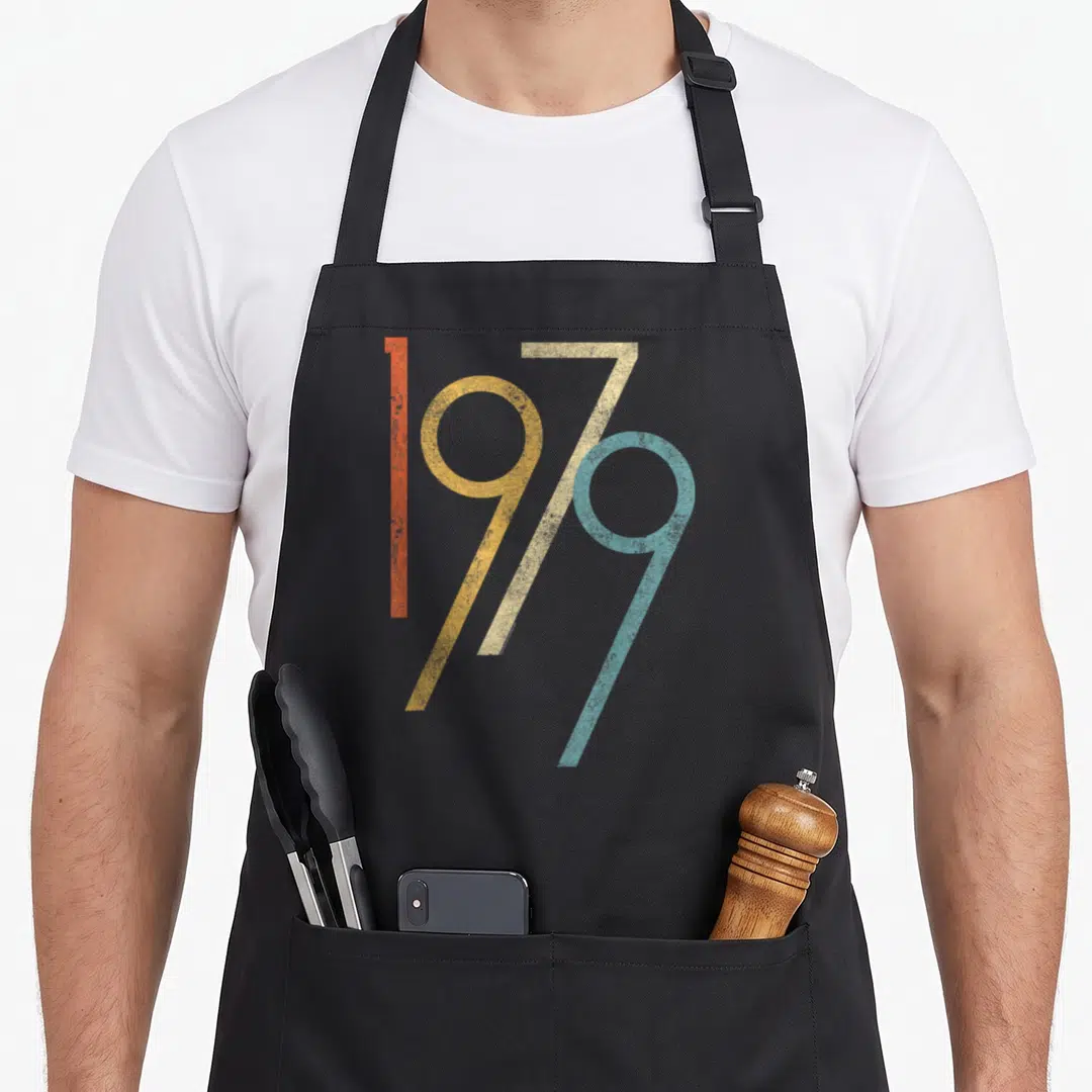 Gen birthday apron - 1979 text in orange yellow beige and blue retro font on black fabric — shown on a black cotton canvas apron against a white background