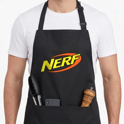 Funny girl birthday apron - Yellow NERF text inside an orange oval swoosh on grey background — shown on a black cotton canvas apron against a white background