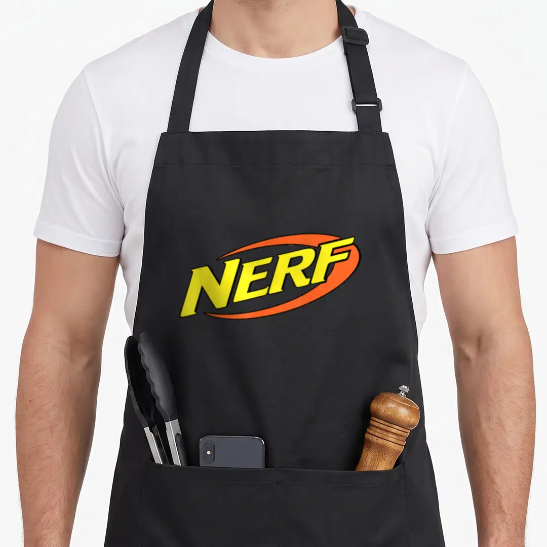 Funny girl birthday apron - Yellow NERF text inside an orange oval swoosh on grey background — shown on a black cotton canvas apron against a white background