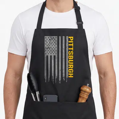 Pittsburgh american flag apron - Vertical grey distressed usa flag with yellow pitsburgh text running alongside stripes — shown on a black cotton canvas apron against a white background