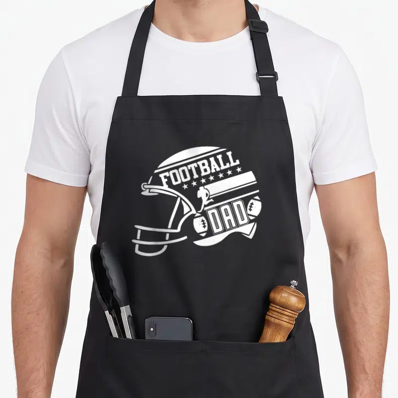 Football father day apron - White football helmet graphic formed by FOOTBALL DAD text and star icons — shown on a black cotton canvas apron against a white background