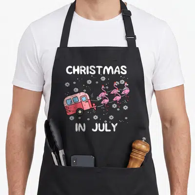 Christmas camp chef apron - Red camper towing five pink flamingos in santa hats under white text reading CHRISTMAS — shown on a black cotton canvas apron against a white background