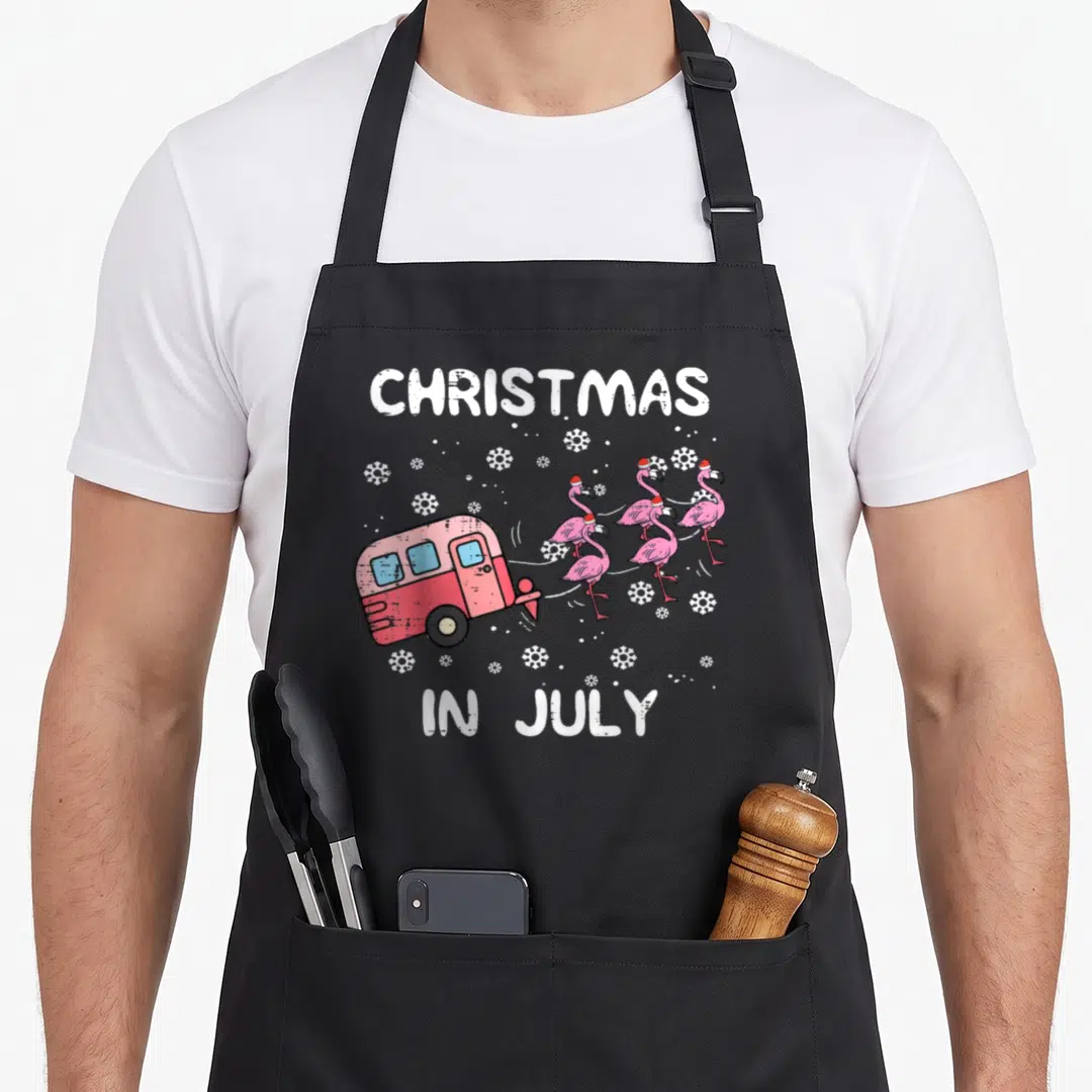 Christmas camp chef apron - Red camper towing five pink flamingos in santa hats under white text reading CHRISTMAS — shown on a black cotton canvas apron against a white background