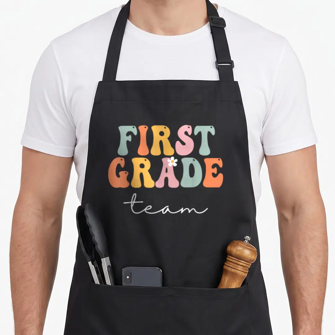 Groovy day grade apron - Bubbly mint orange yellow pink and white text reading FIRST GRADE team with a small flower — shown on a black cotton canvas apron against a white background
